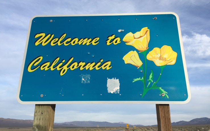 A blue road sign that says Welcome to California in script, alongside a picture of a California Golden Poppy. Behind the sign is a highway, leading to mountains and a cloudy, but bright horizon.
